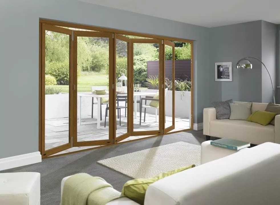 A living room with white sofas looking through a set of half open brown bifold doors looking out towards a bright garden with chairs and a dining table